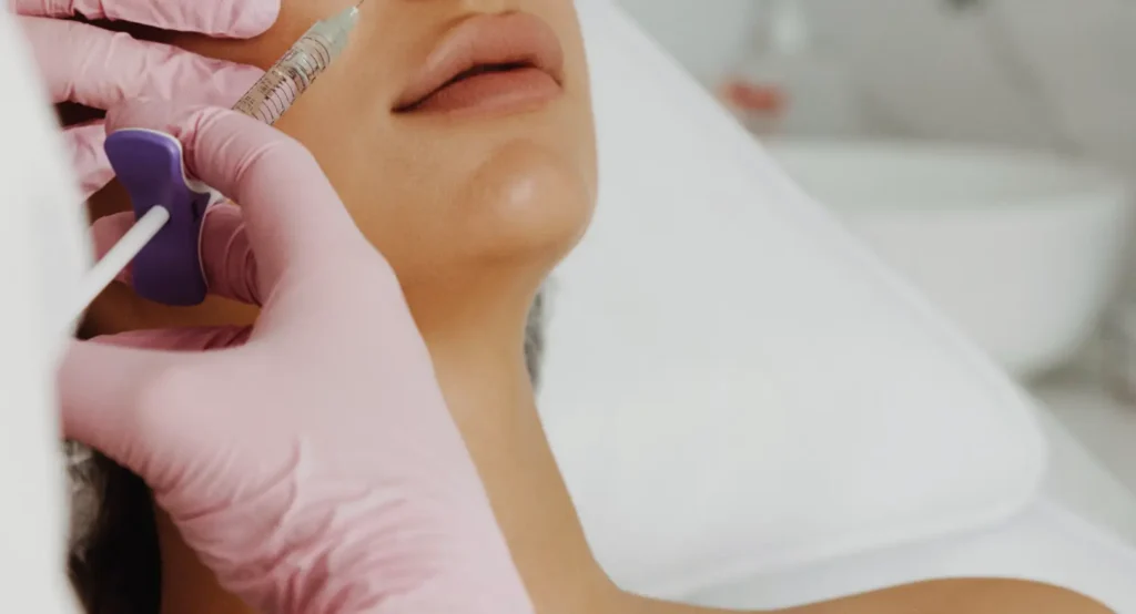 Practitioner wearing pink gloves holds a syringe near a woman's lower cheek and mouth.