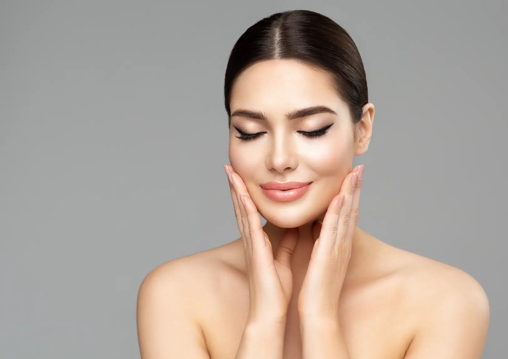 Woman with closed eyes gently touches both sides of her face against a gray background.